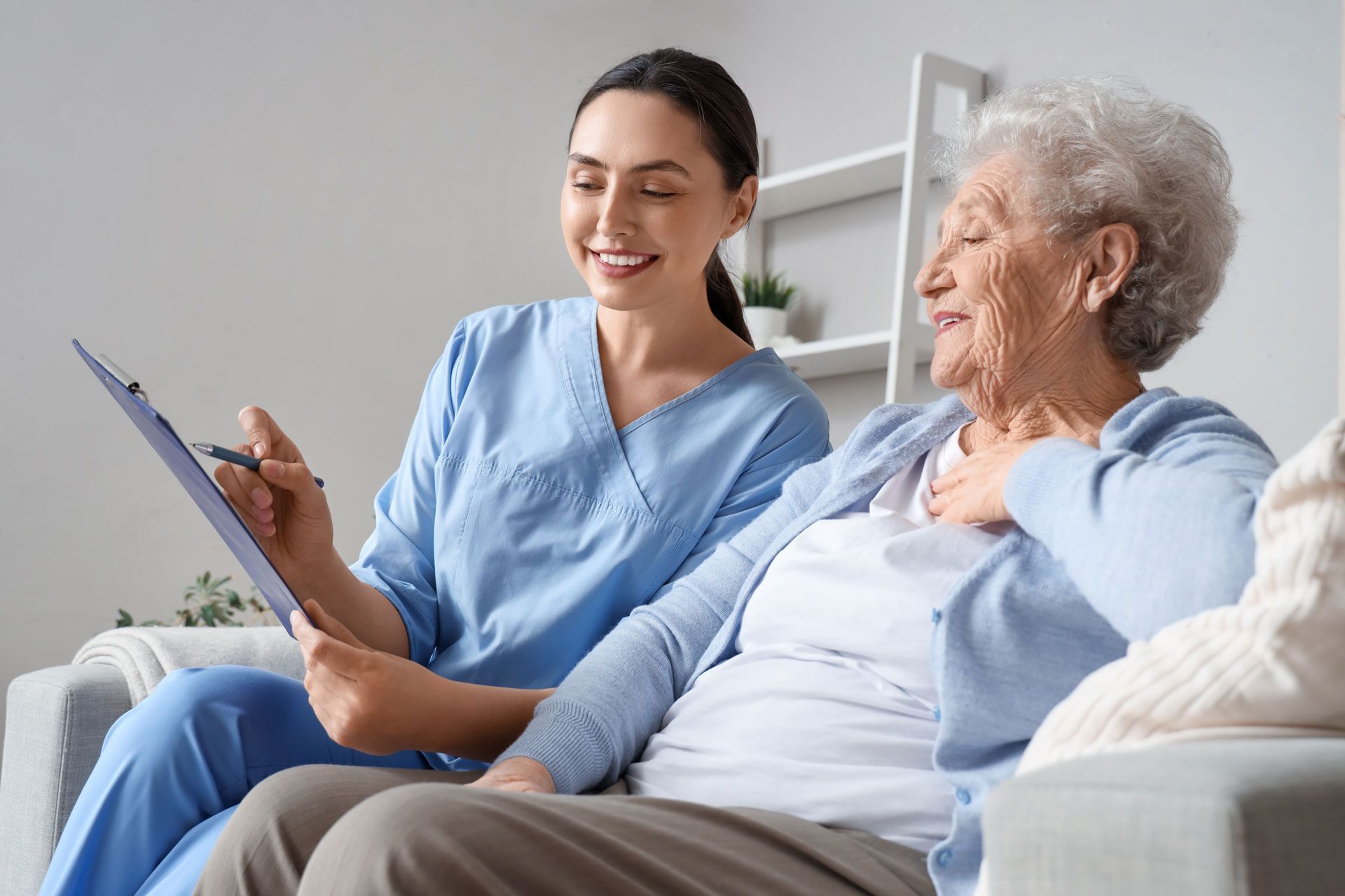 A caregiver in blue scrubs points to a clipboard, smiling at an older woman seated next to her on a sofa.