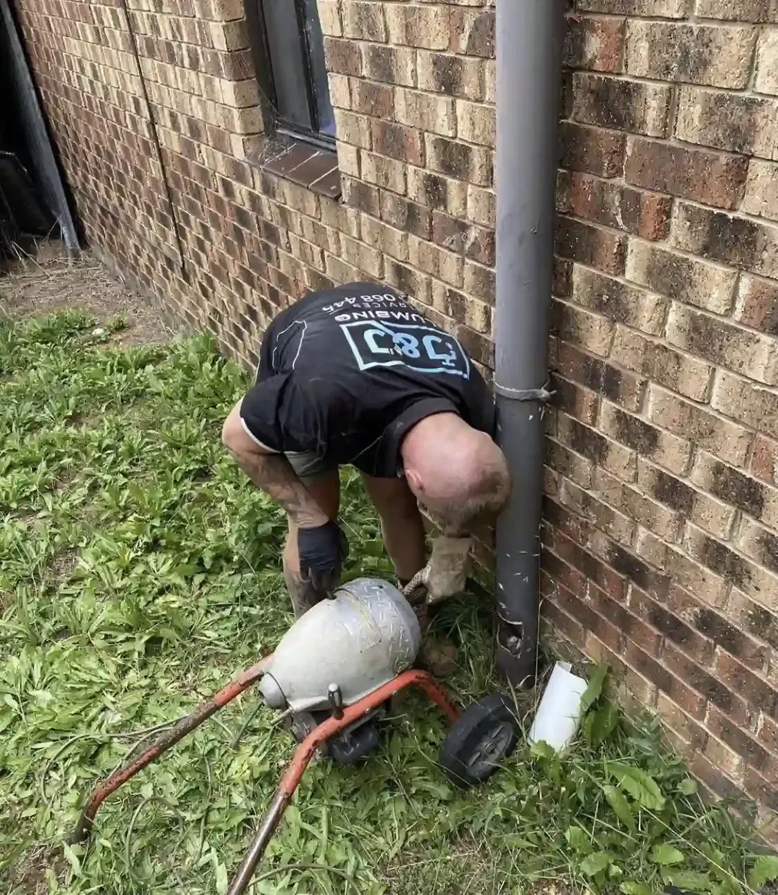Person crouching beside a brick wall, working on a metal tank and hoses on a wheeled cart.
