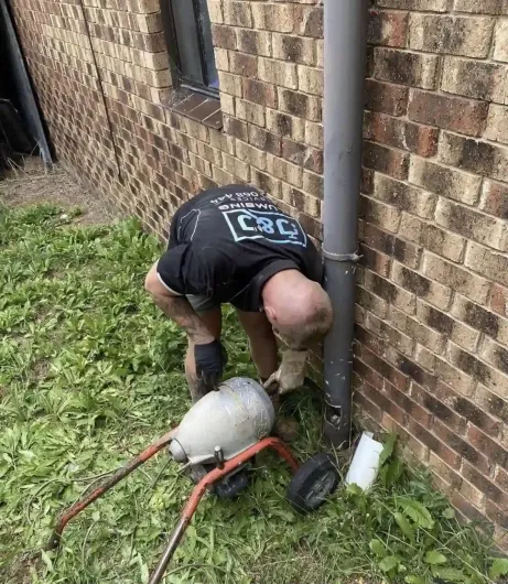 Person bending over a tank and hose setup beside a brick wall and downspout in a yard