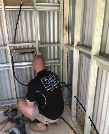 Plumber kneeling inside a wall frame, inspecting pipes in an unfinished room.