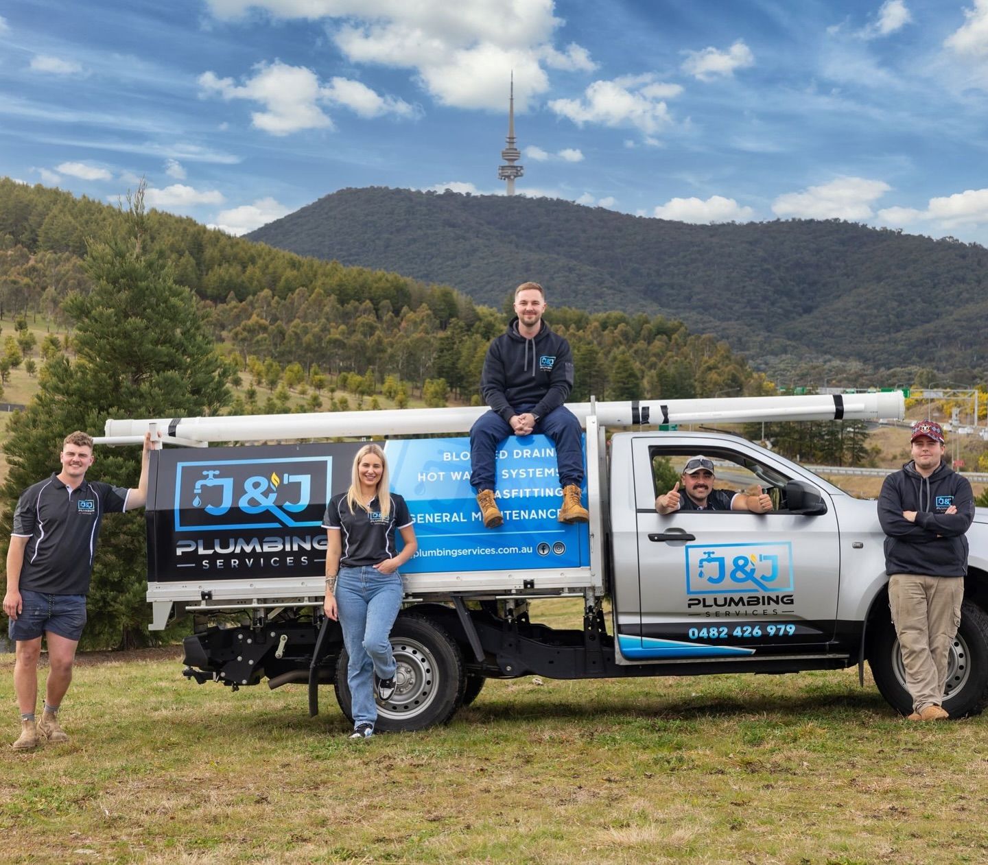 Four people posing with a blue utility truck in a grassy field with mountains and a cloudy sky behind them.