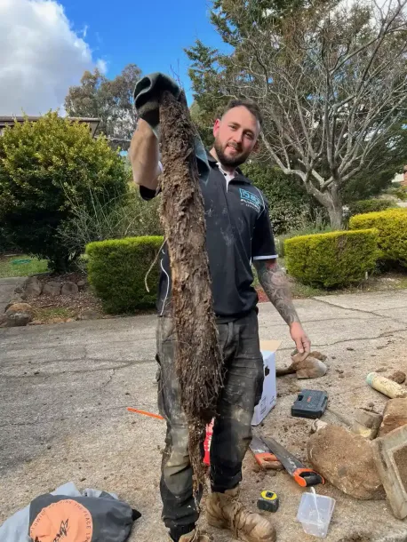 Man holding a large catfish outdoors on a driveway, with yard tools and a stone pile nearby.