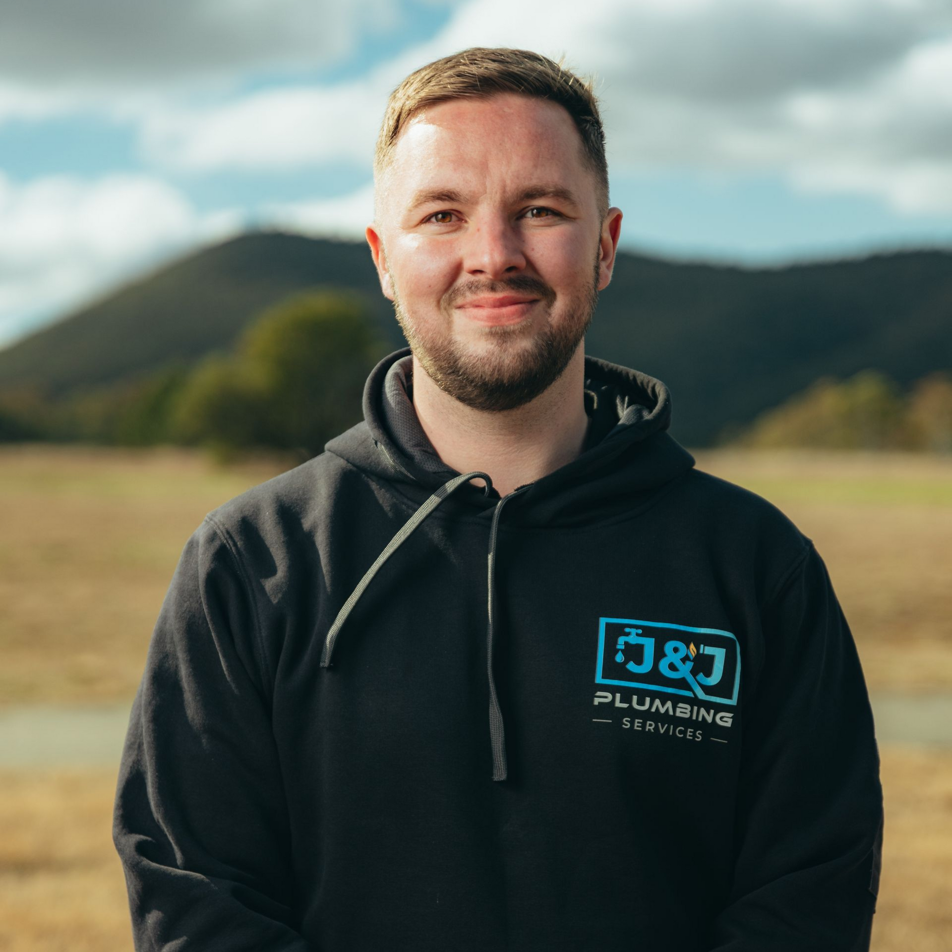 Man in a black hoodie standing outdoors with mountains and cloudy sky in the background