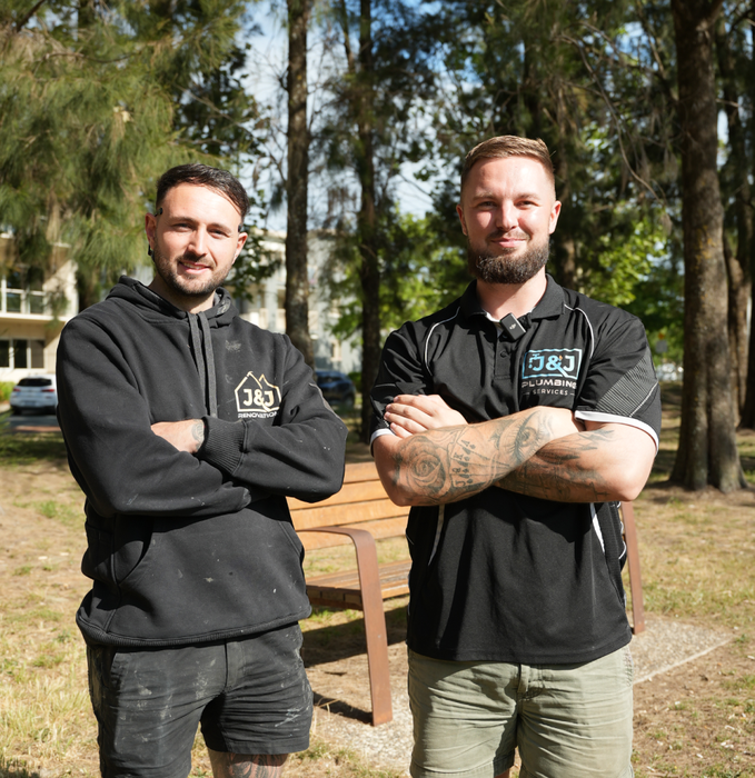 Two men with crossed arms standing outdoors in a sunny park-like setting.