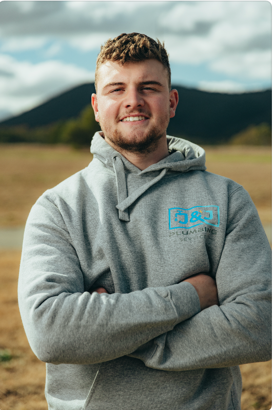 Person in a gray hoodie standing outdoors with arms crossed, mountains blurred in the background