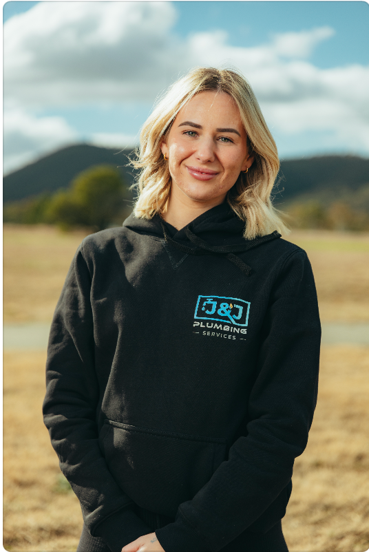 Person in a black hoodie stands outdoors in a field with mountains and a cloudy sky.
