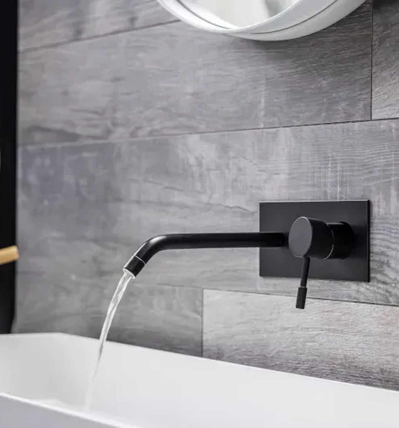Wall-mounted black faucet filling a white bathtub against gray tile.