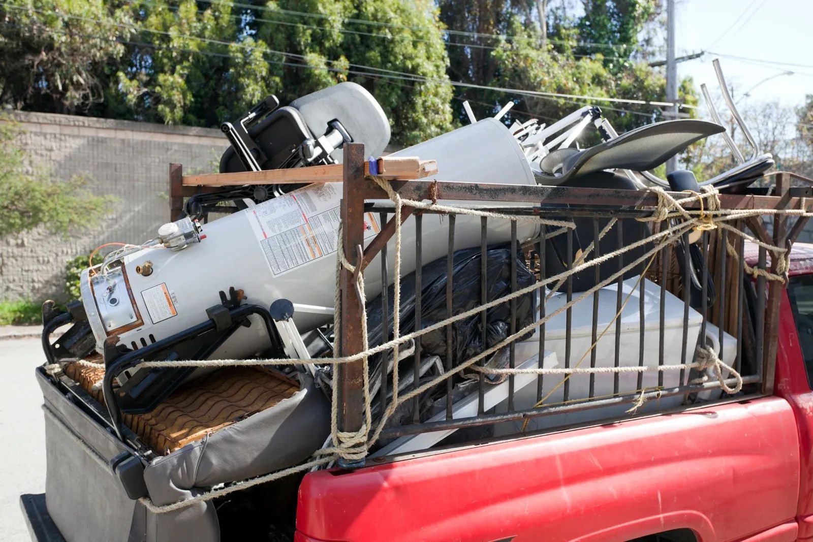 Loaded red pickup truck piled high with scrap metal and old appliances by a roadside wall