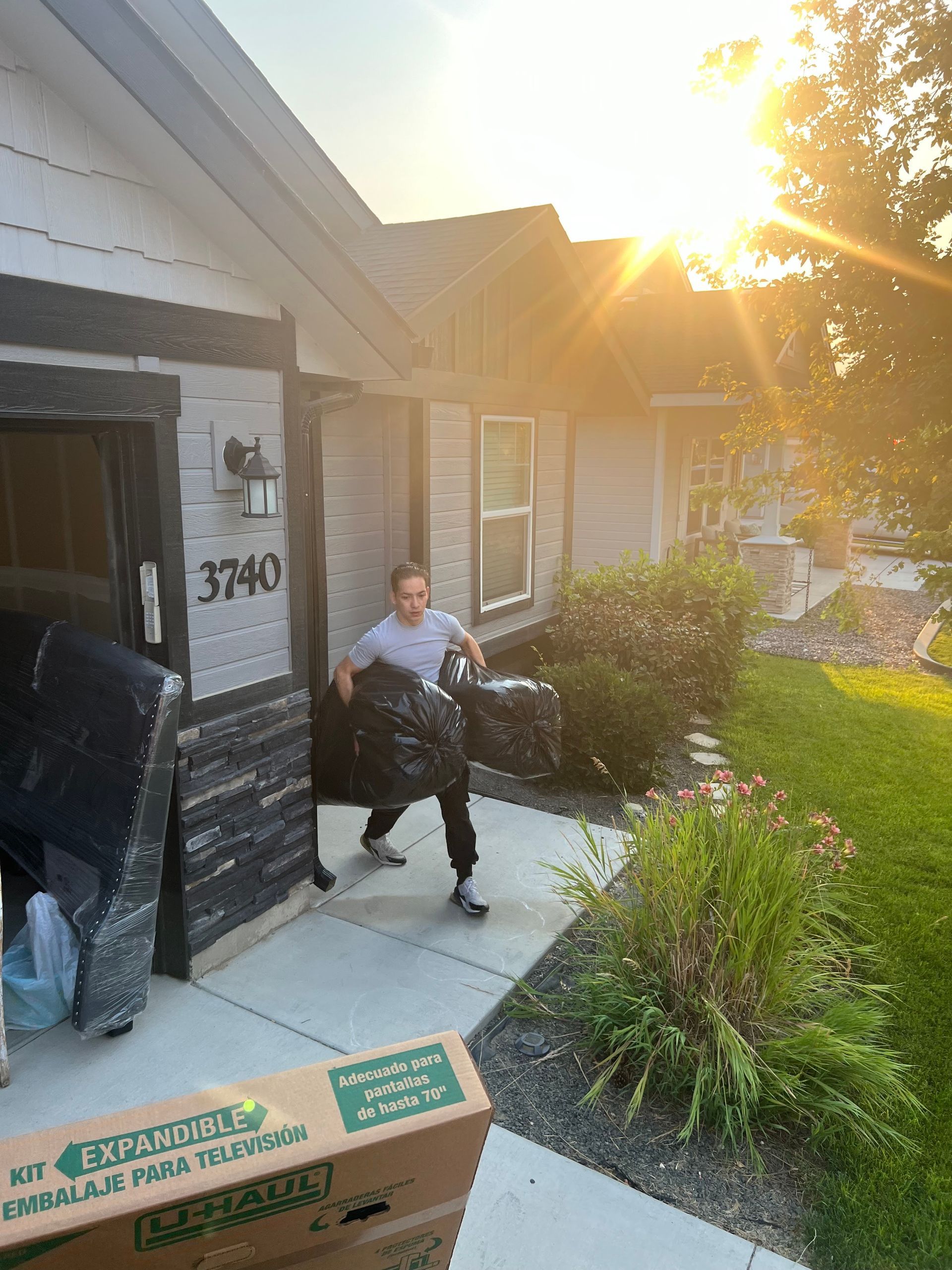 Person carrying trash bags outside a house with sunlight; boxes and furniture are visible.