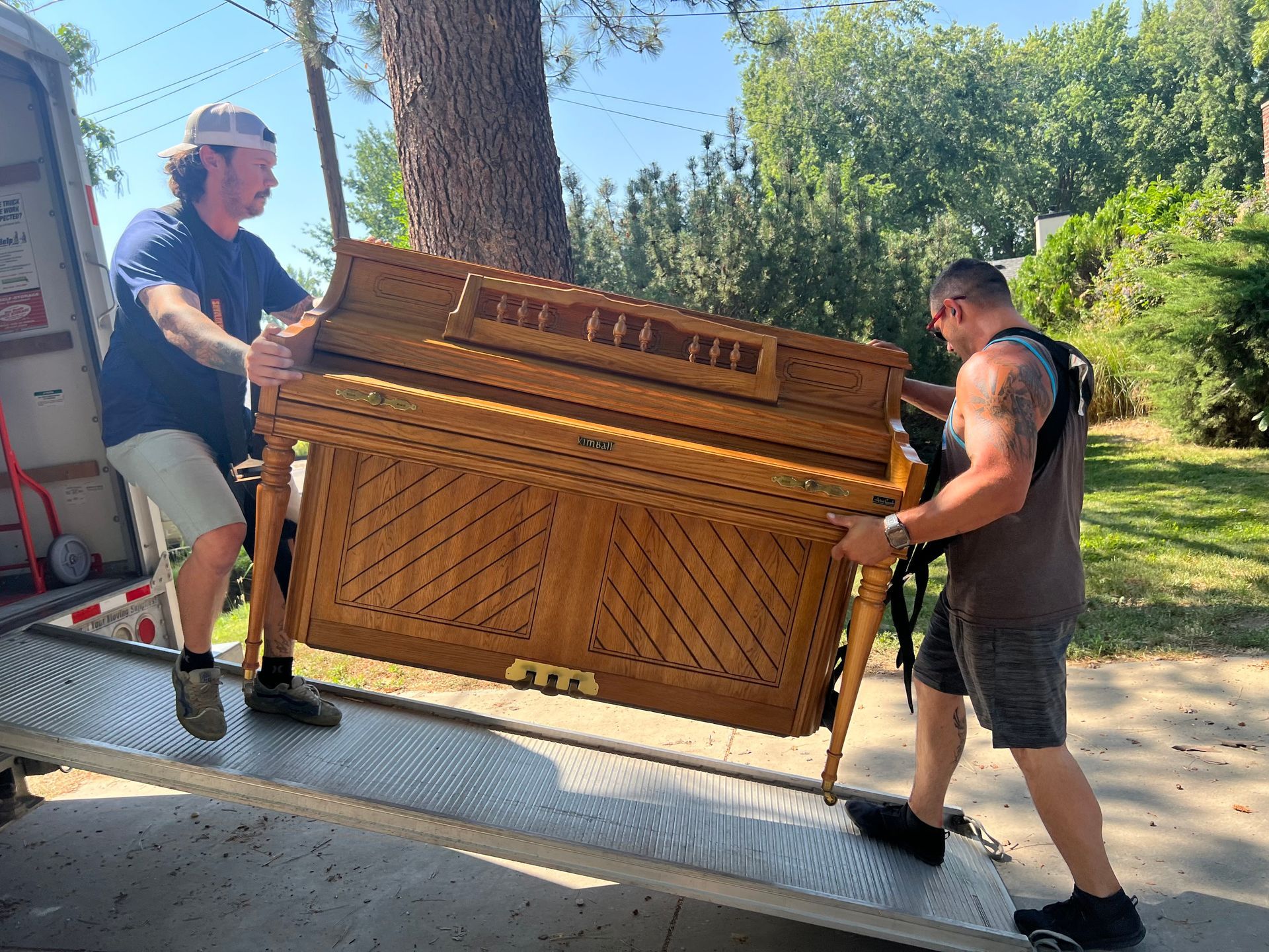 Two men carrying a wooden piano up a ramp into a truck.