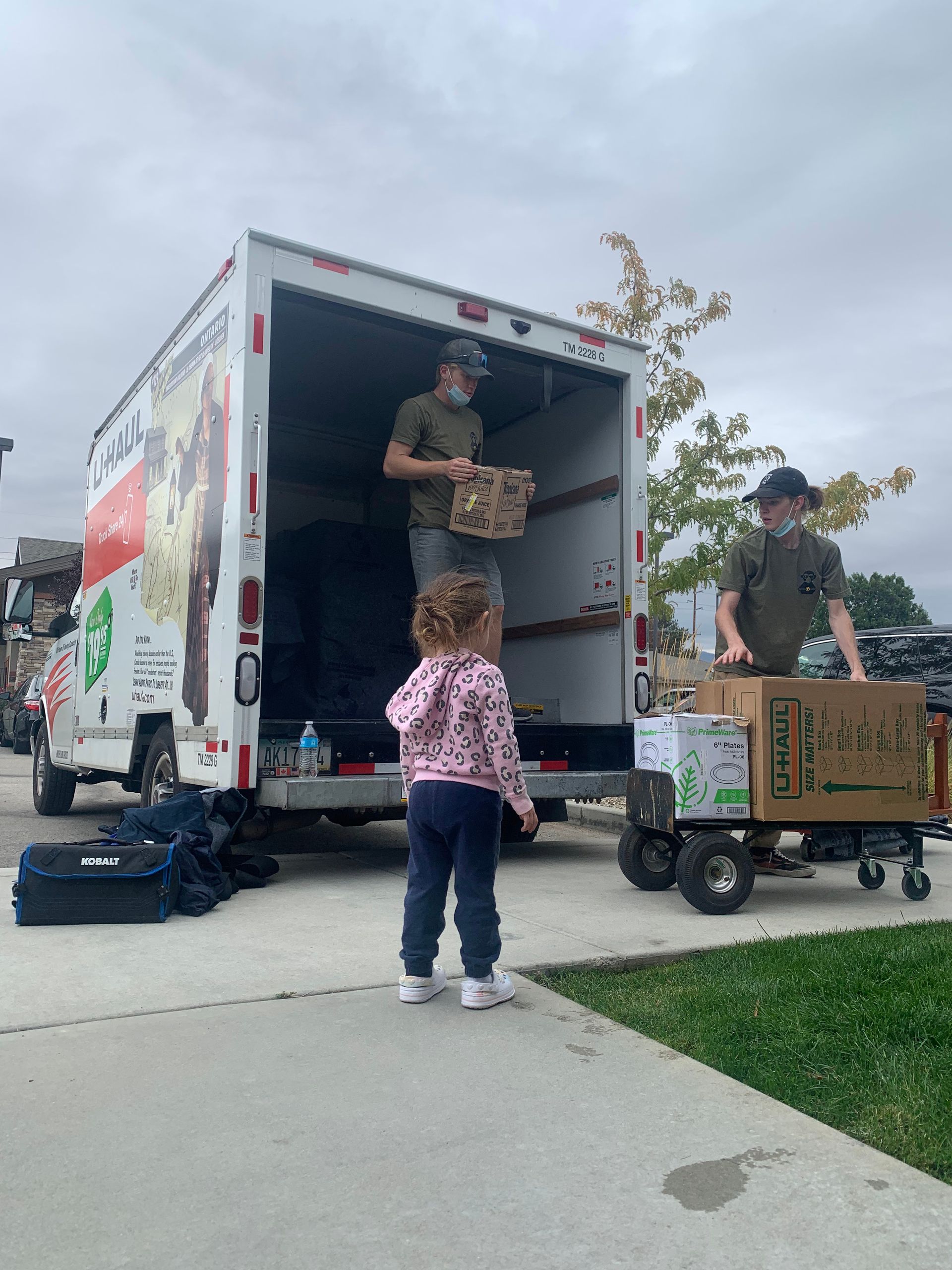 Two people unloading boxes from a truck, child watches. Truck has a logo. Outdoors.