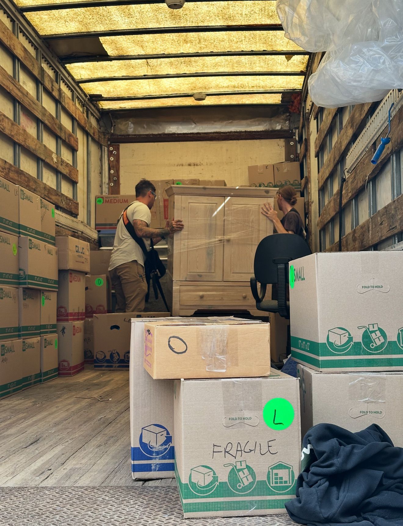 Two people loading a large wooden cabinet into the back of a moving truck filled with stacked cardboard boxes.