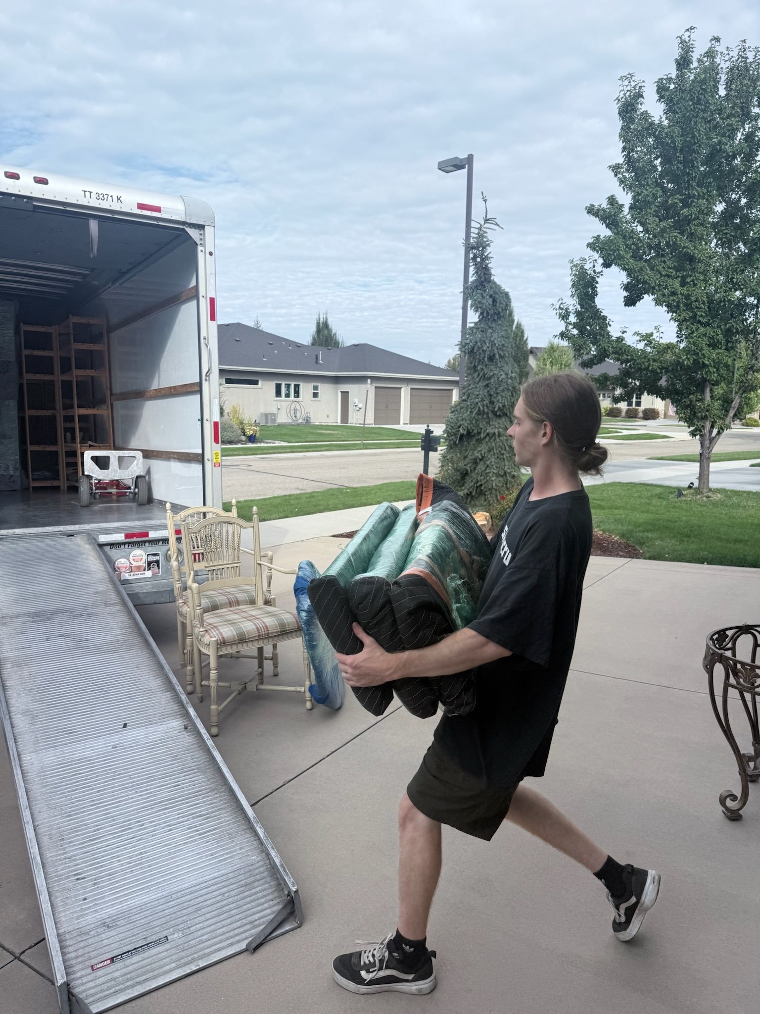 Man carrying a large, dark object from a moving truck onto a driveway.  A ramp is in use.