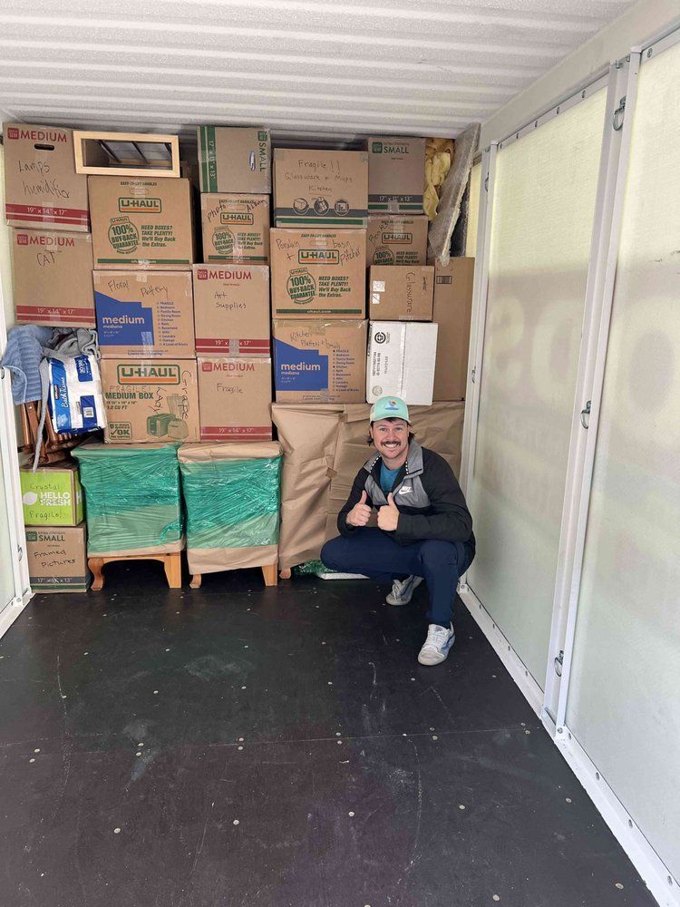 Man squats in a storage container filled with stacked moving boxes, giving a thumbs-up.