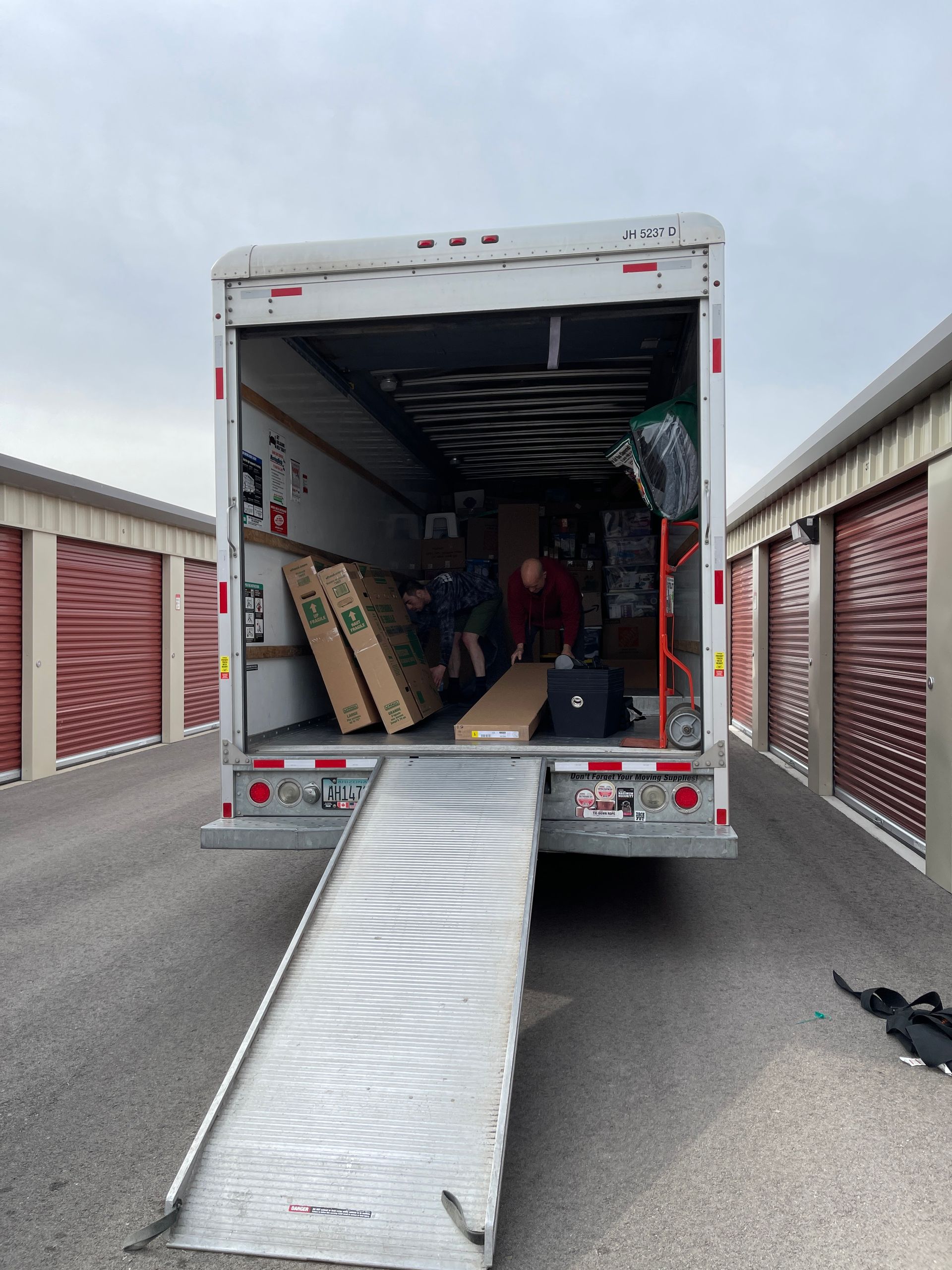 A moving truck with ramp open, loaded with furniture, parked between storage units.
