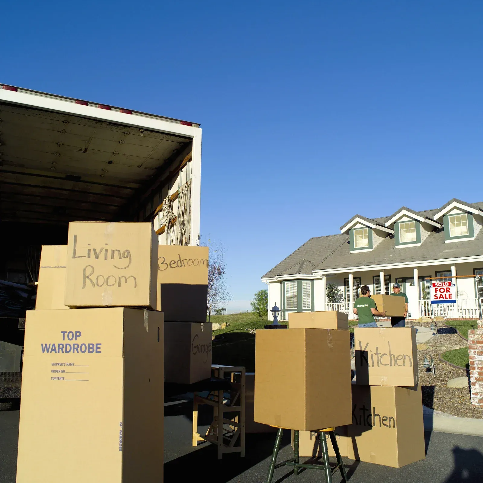 Moving boxes stacked by a truck in front of a house; people unloading.