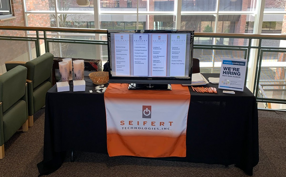 Seifert Technologies info table, with cups, business cards, pencils, monitor, and a display card with