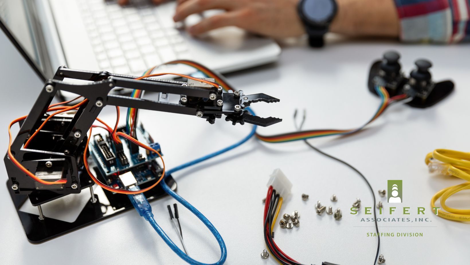 Someone typing at a laptop. Connected to the laptop ports is a small robotic arm.