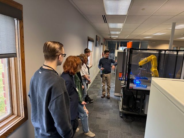Students gathered around a robot arm. A man standing next to and is presumably talking about the robot.