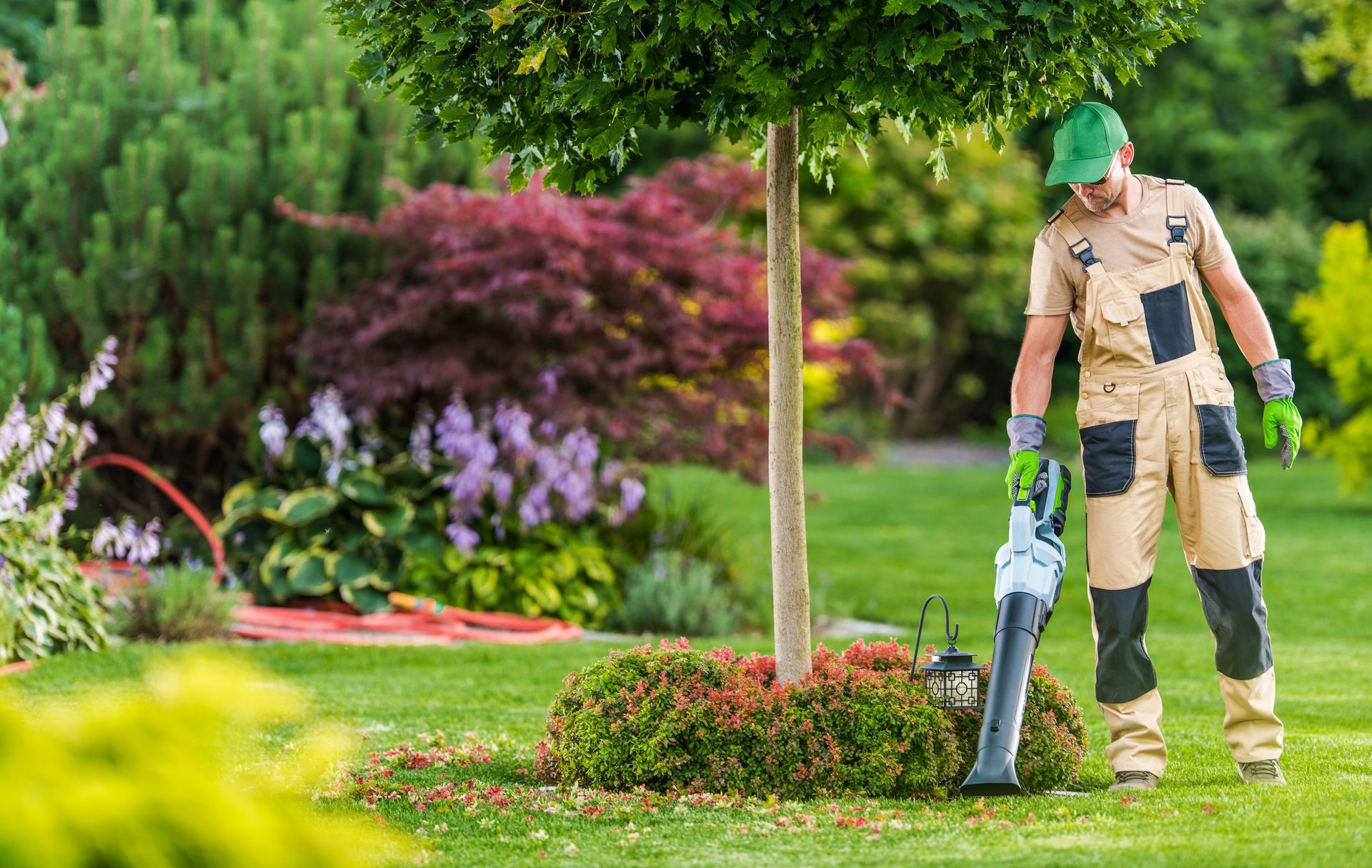 Gardener in beige workwear using a leaf blower in a lush green park with flowers and shrubs