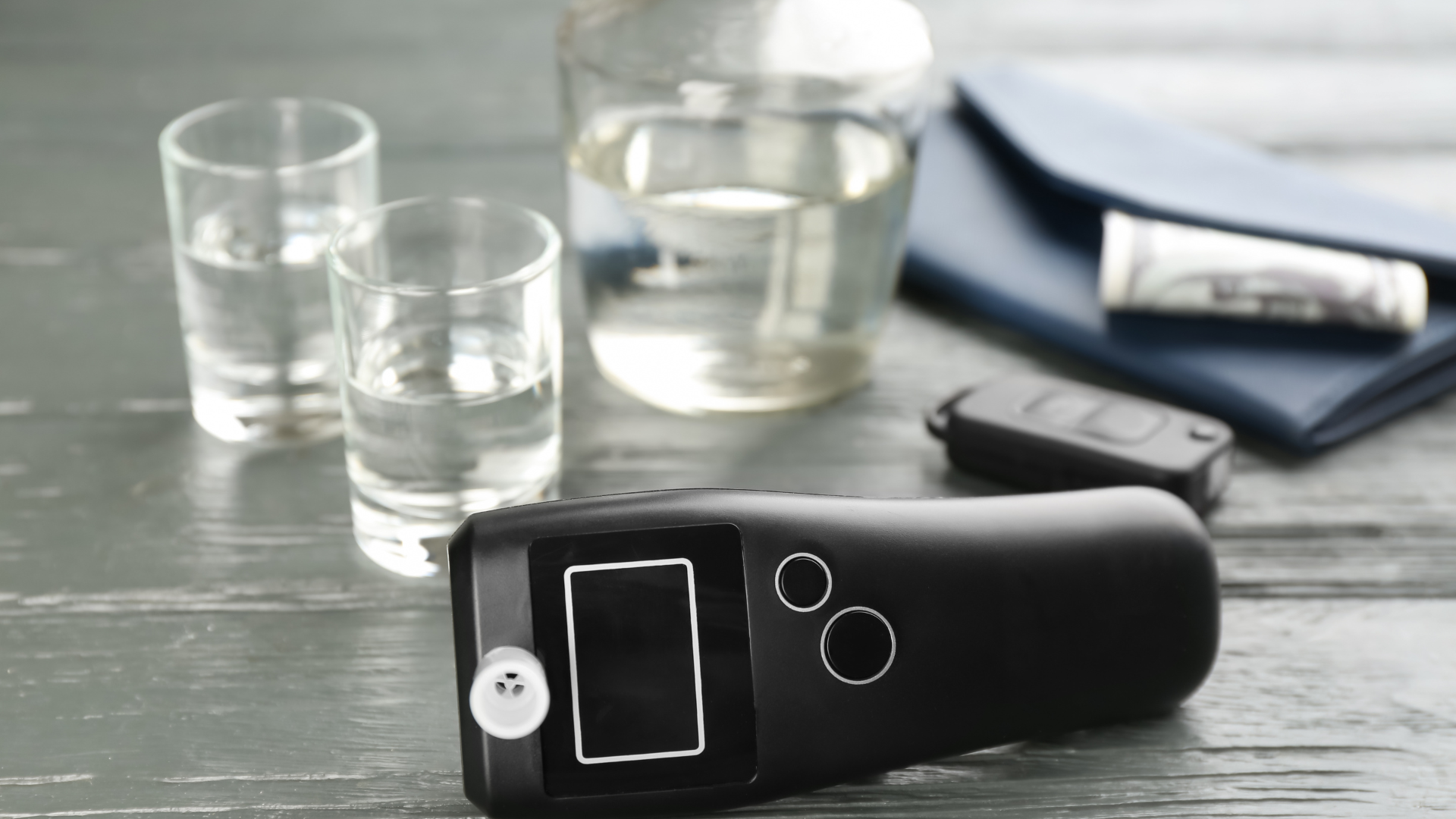 A digital breathalyzer on a wooden surface with two shot glasses, a glass of water, and a wallet in the background.
