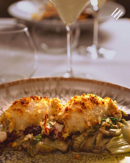 Un piatto di pasta al forno con formaggio, servito su un piatto bianco al tavolo di un ristorante, con un bicchiere di vino sullo sfondo.