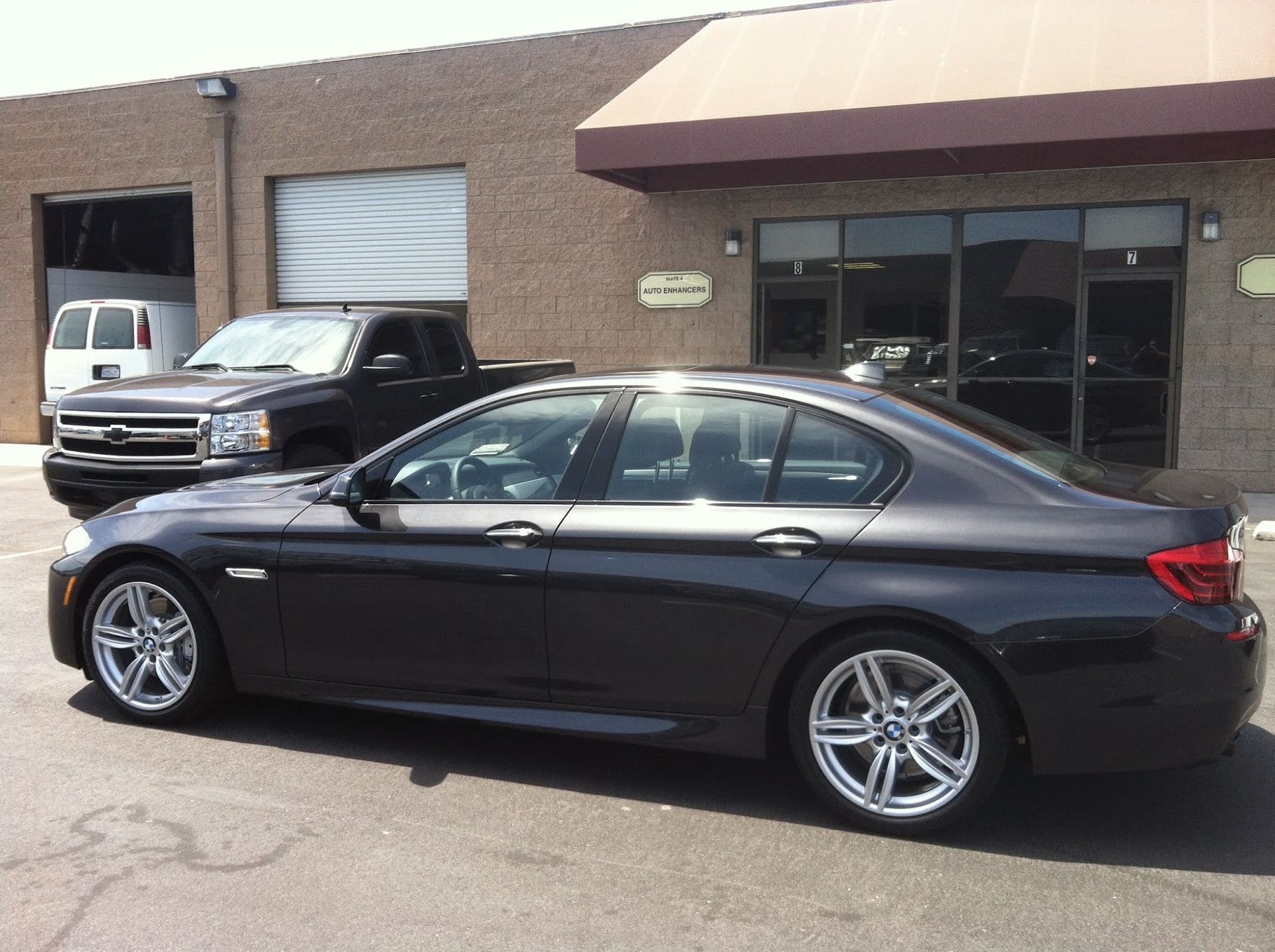 Dark gray BMW sedan parked outside a brick building with a pickup truck and white van visible.