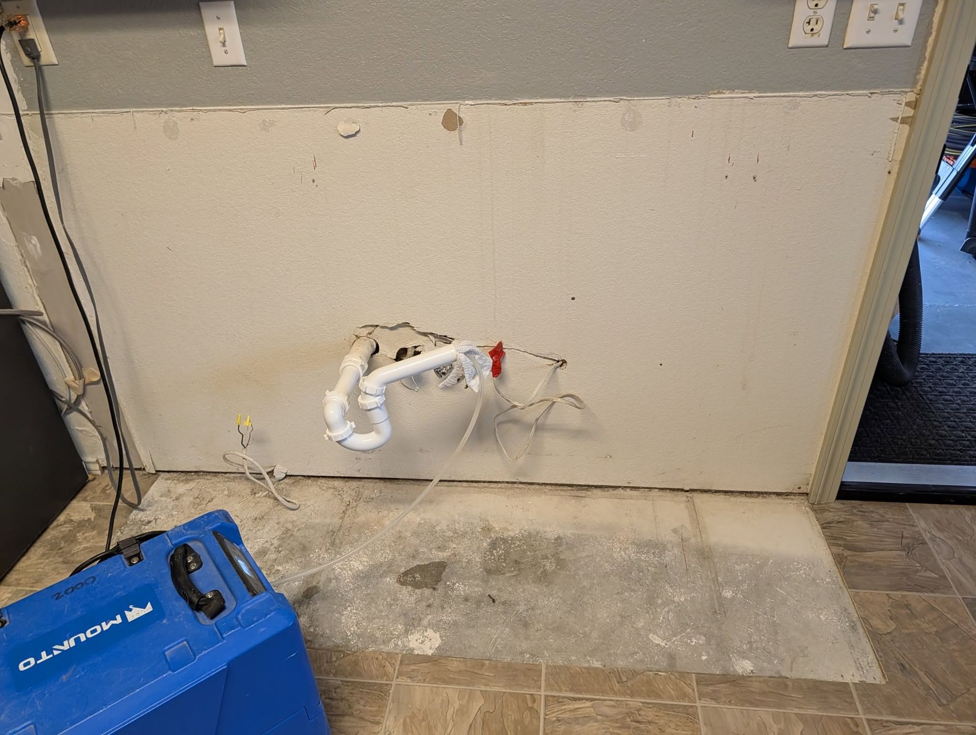 Damaged wall with exposed plumbing pipe and debris on the floor beside a blue machine