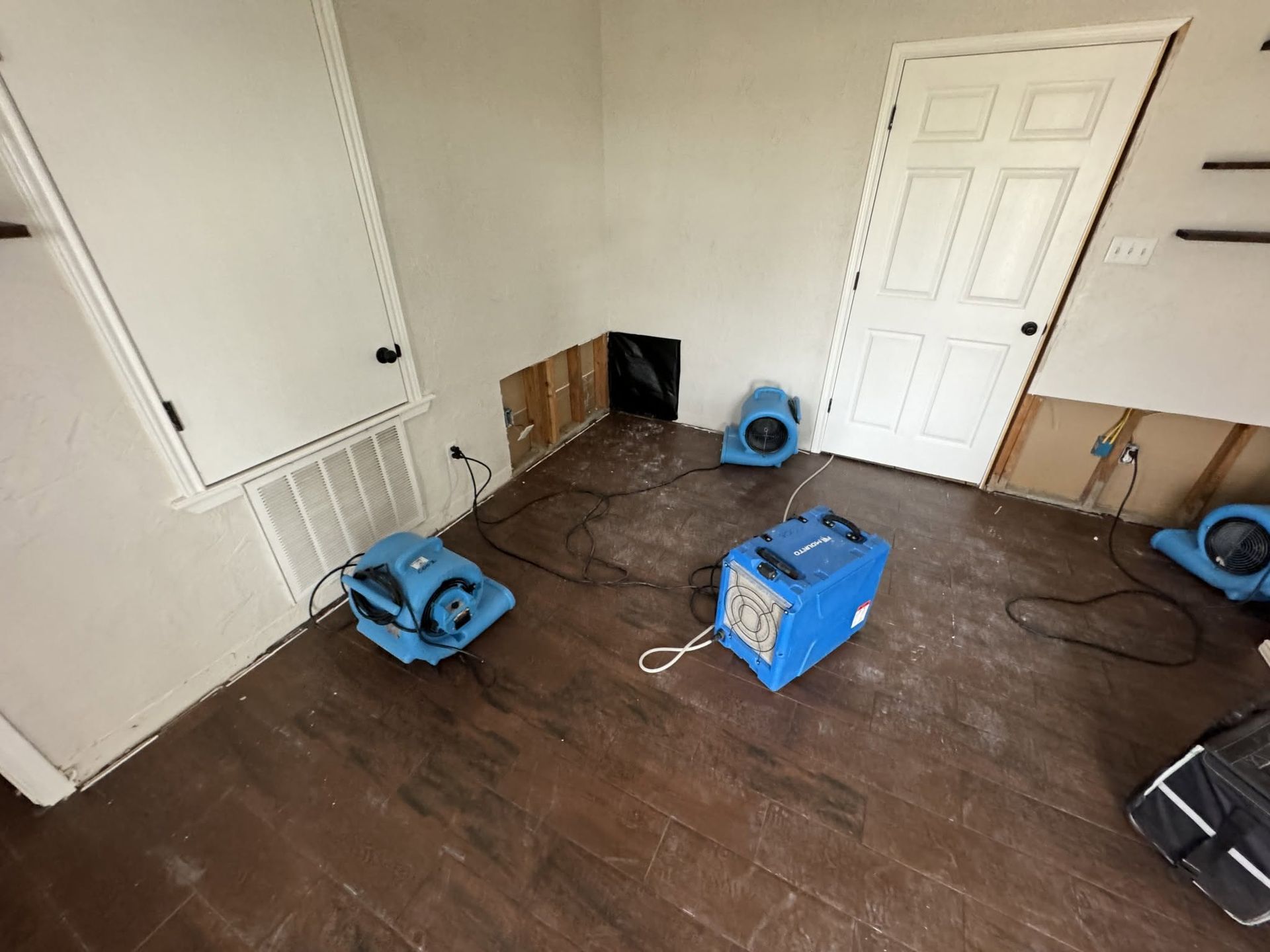 An empty room with brown flooring, a white door, and several blue fans and cords on the floor.