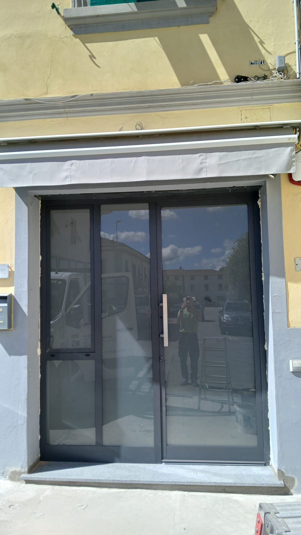 Vetrine chiuse con telaio grigio e tenda da sole su un edificio giallo.