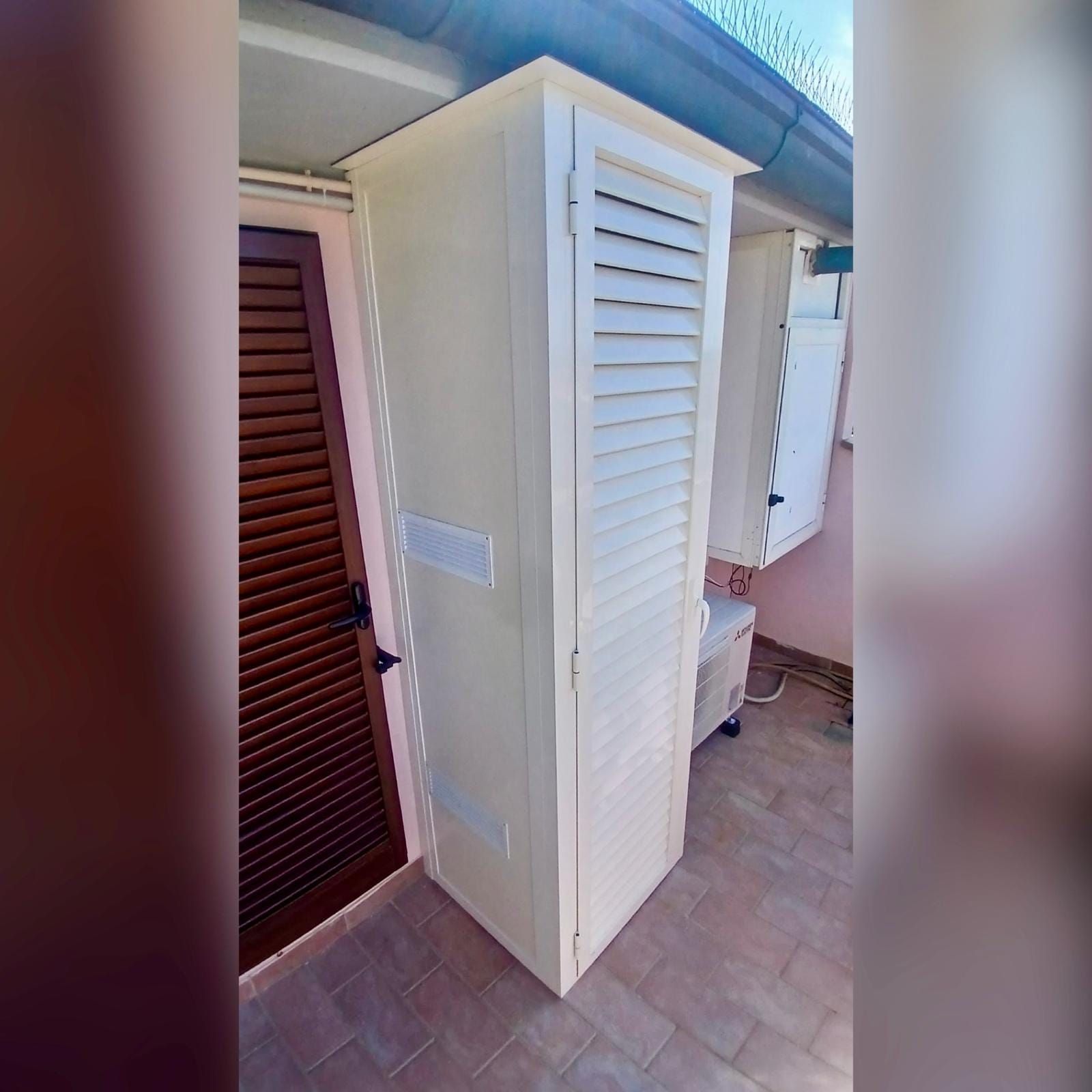 Mobiletto da esterno bianco con ante a persiana su un patio piastrellato accanto a un muro e porta con persiana marrone