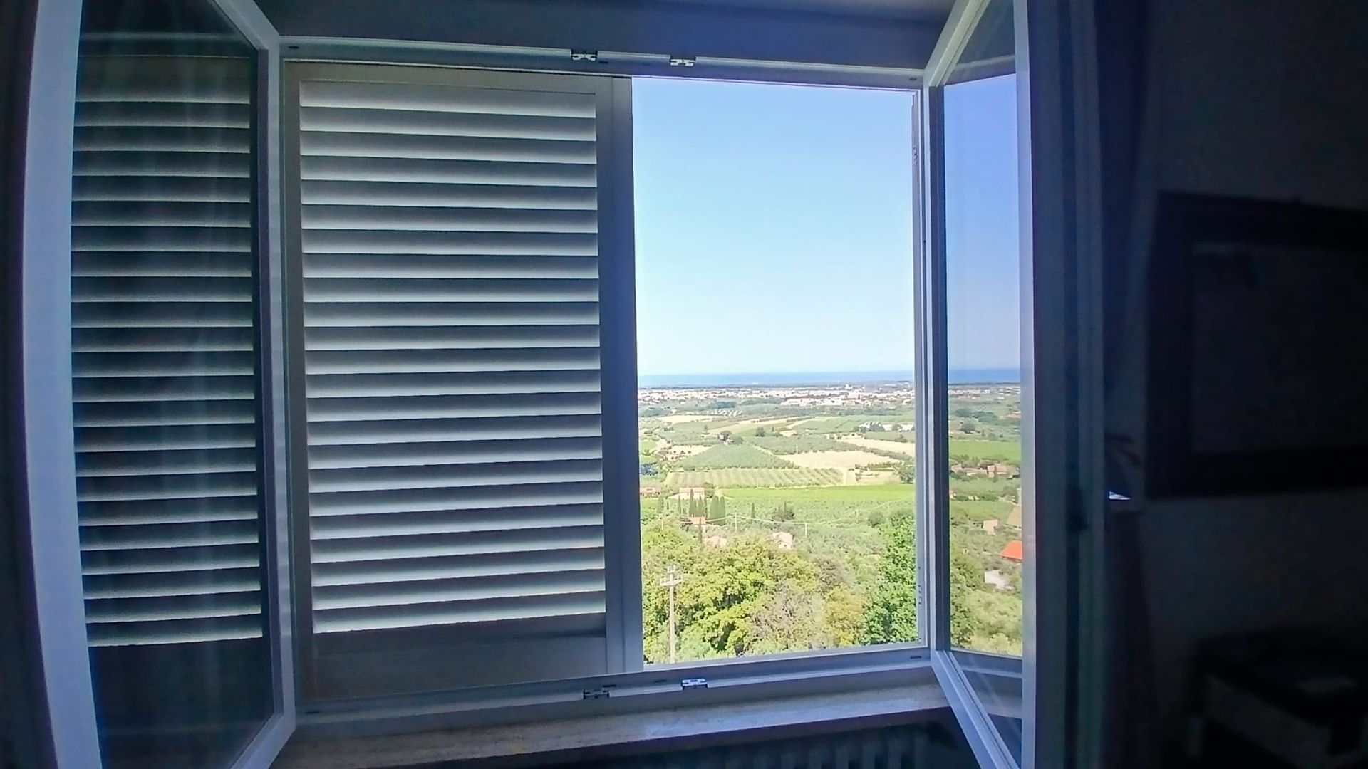 Vista attraverso una finestra con persiane bianche su un paesaggio verde brillante e un cielo azzurro.