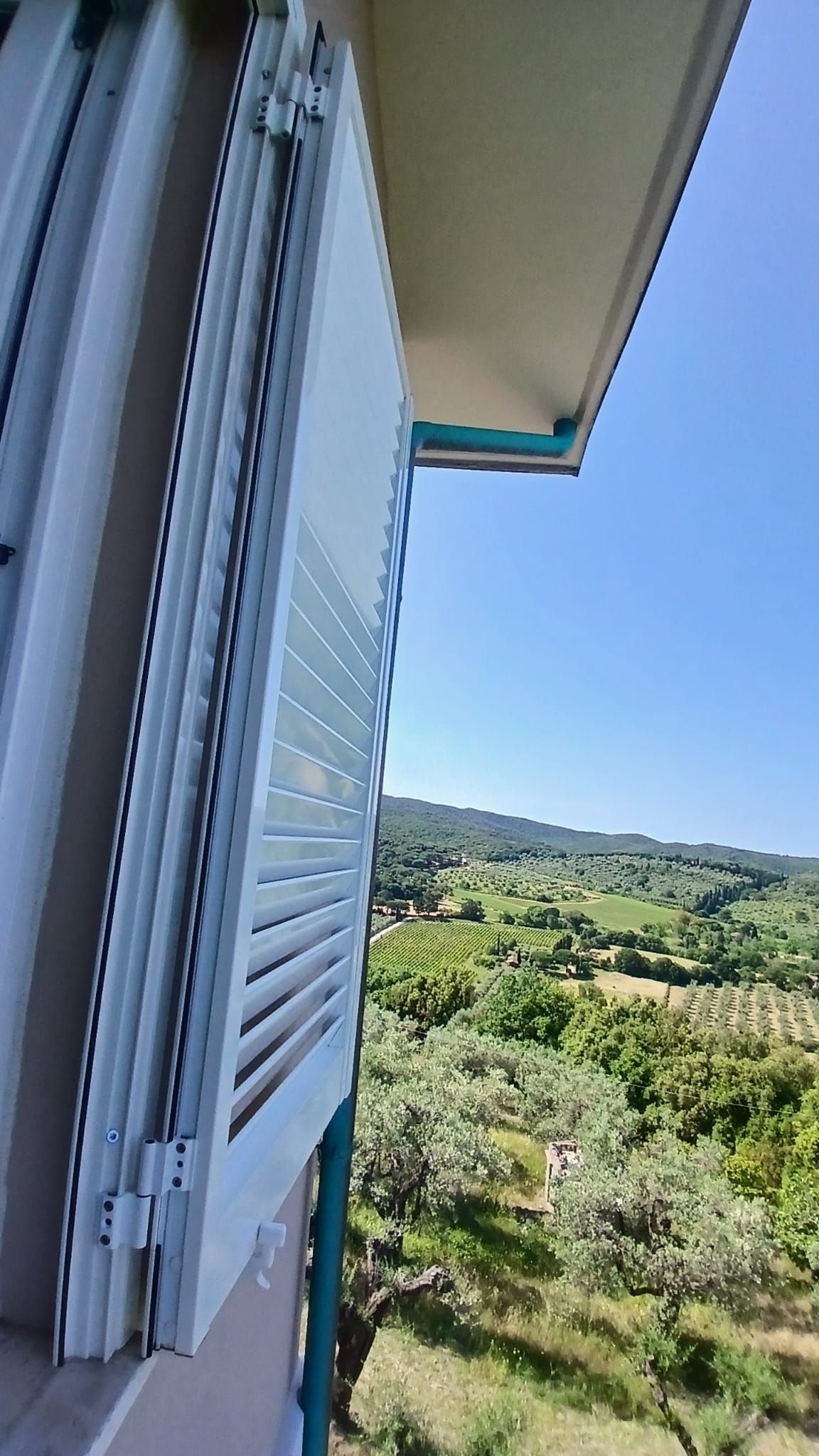 Persiane aperte che si affacciano su una verde vallata illuminata dal sole e un cielo azzurro.