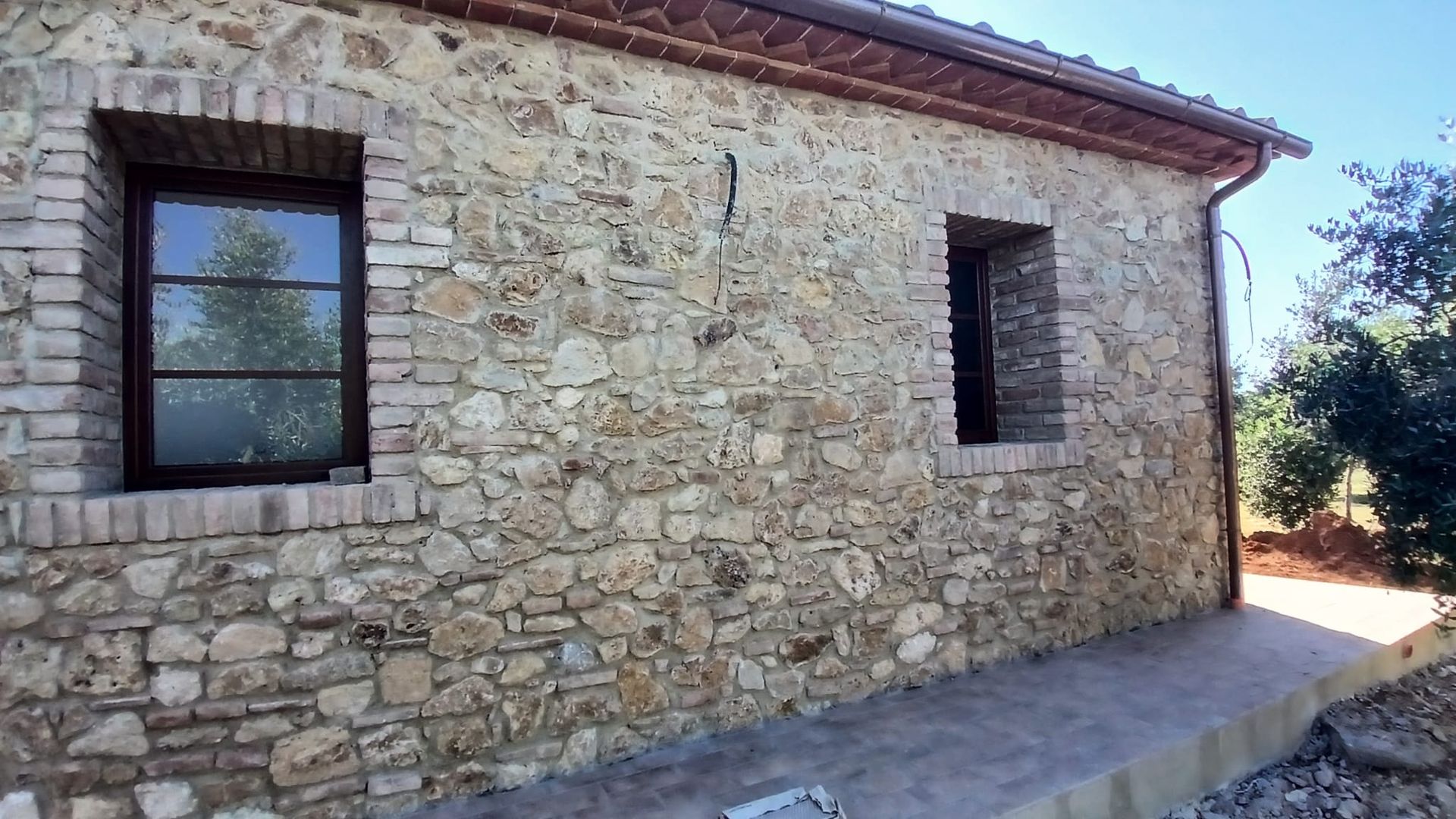 Esterno di una casa in pietra con due finestre e tetto di tegole, in piena luce del sole.