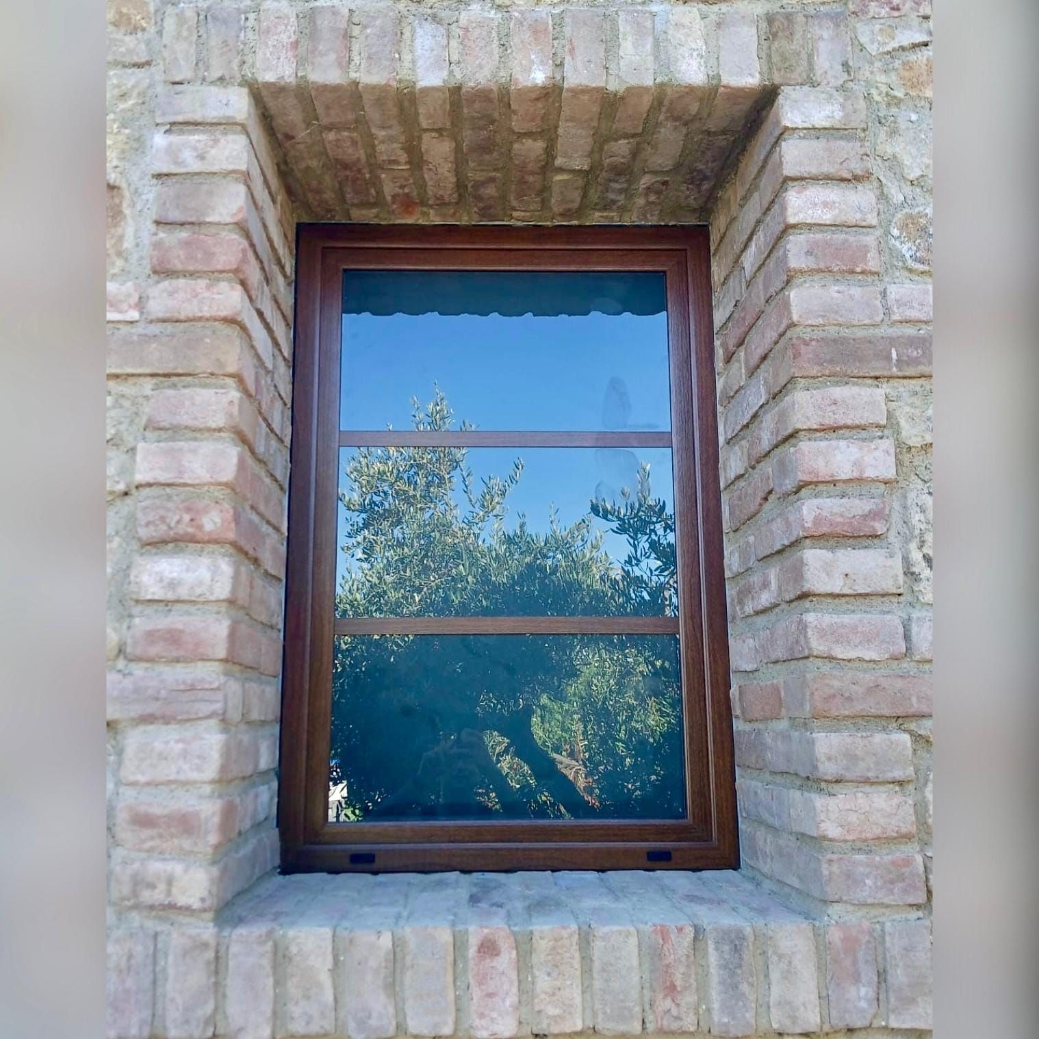 Finestra con cornice in legno incastonata in un muro di pietra chiara, che riflette gli alberi e il cielo azzurro.