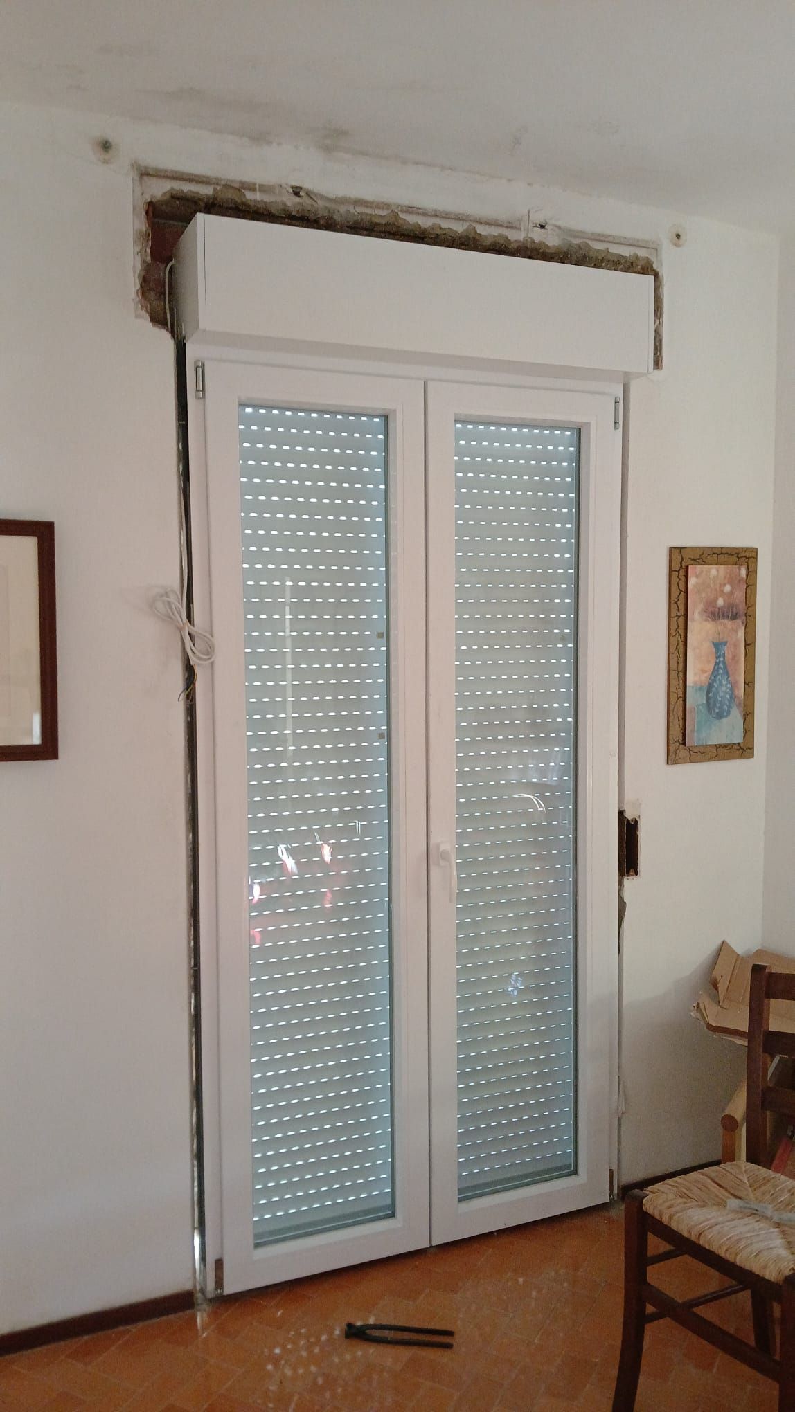Doppie porte a vetri bianche sotto un'apertura in un muro grezzo e non rifinito, in una stanza semplice con una sedia e un quadro incorniciato.