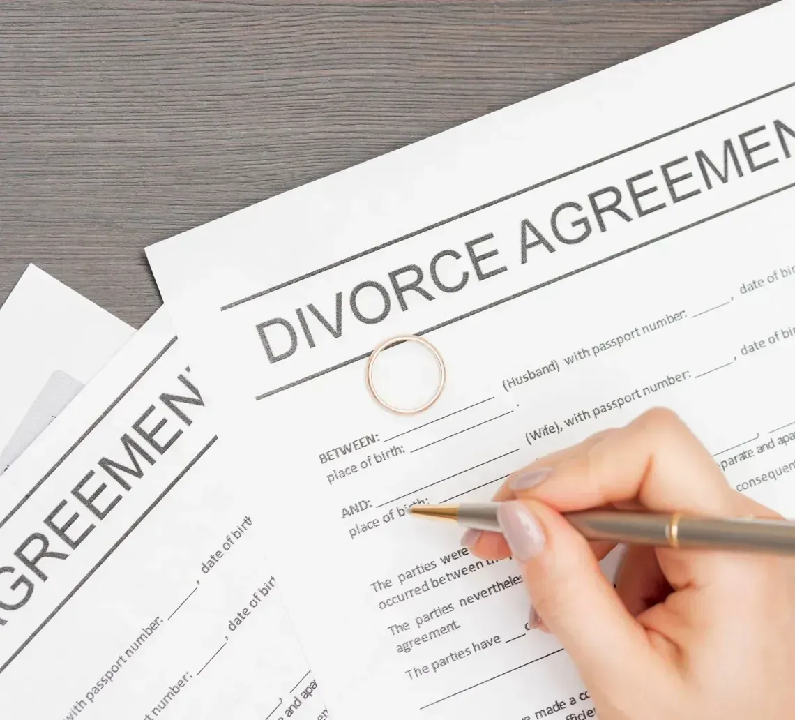 A person's hand holding a pen over a divorce agreement form on a wooden table, with a wedding ring resting on the paper.