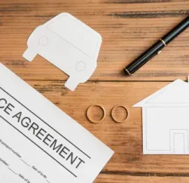 A document titled Divorce Agreement, a pen, two wedding rings, and paper cutouts of a house and car on a wooden table.