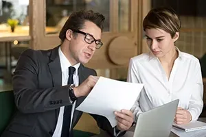 A professional in a suit and glasses holds a document while discussing it with a colleague at a laptop in an office.