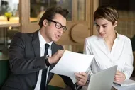 A professional in a suit and glasses holds a document while discussing it with a colleague at a laptop in an office.