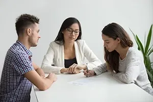 Two people sit at a table across from a professional, who watches as one person signs a document.