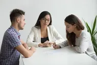 Two people sit at a table across from a professional, who watches as one person signs a document.