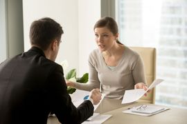 A professional in a suit and a person in a beige top look at documents during a serious discussion in a bright office.