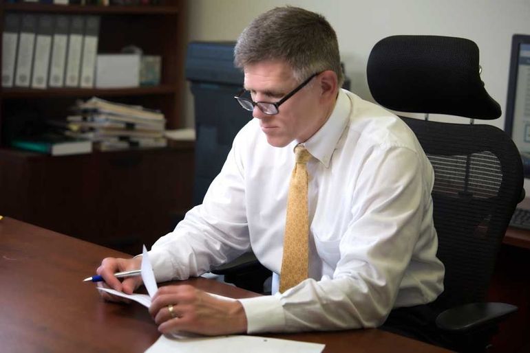 A person wearing a white shirt and gold tie sits at a desk, looking down and working with papers.