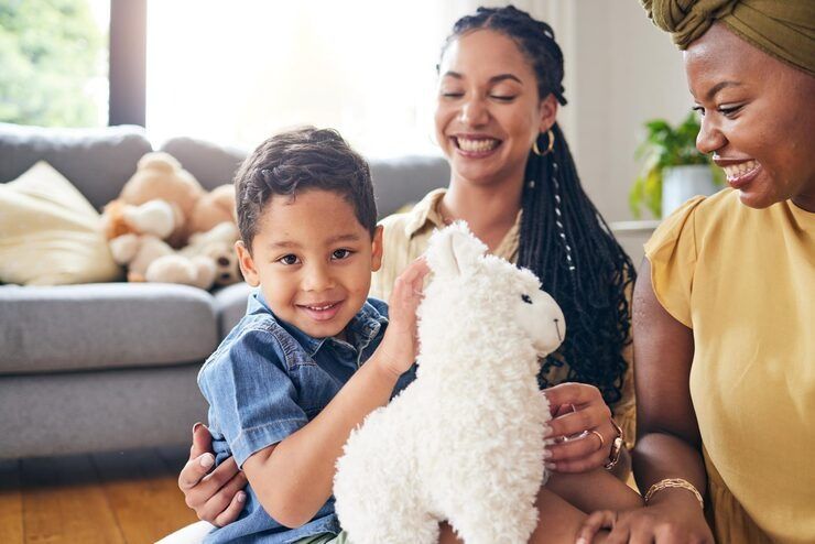 Two people sit on a floor in a living room, smiling as a child holds a plush white llama toy.