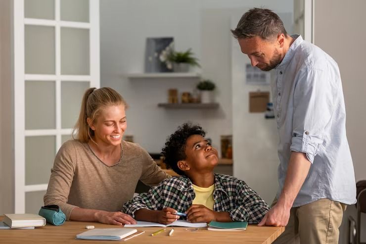 A student sits at a table writing in a notebook while a smiling adult watches, and another adult stands looking down.