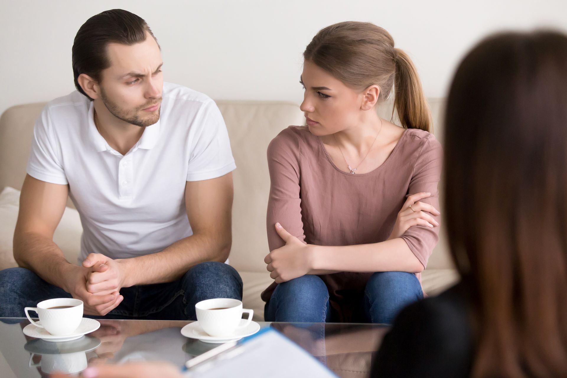 A couple sits on a couch facing a professional during a counseling session, appearing to discuss a serious topic.