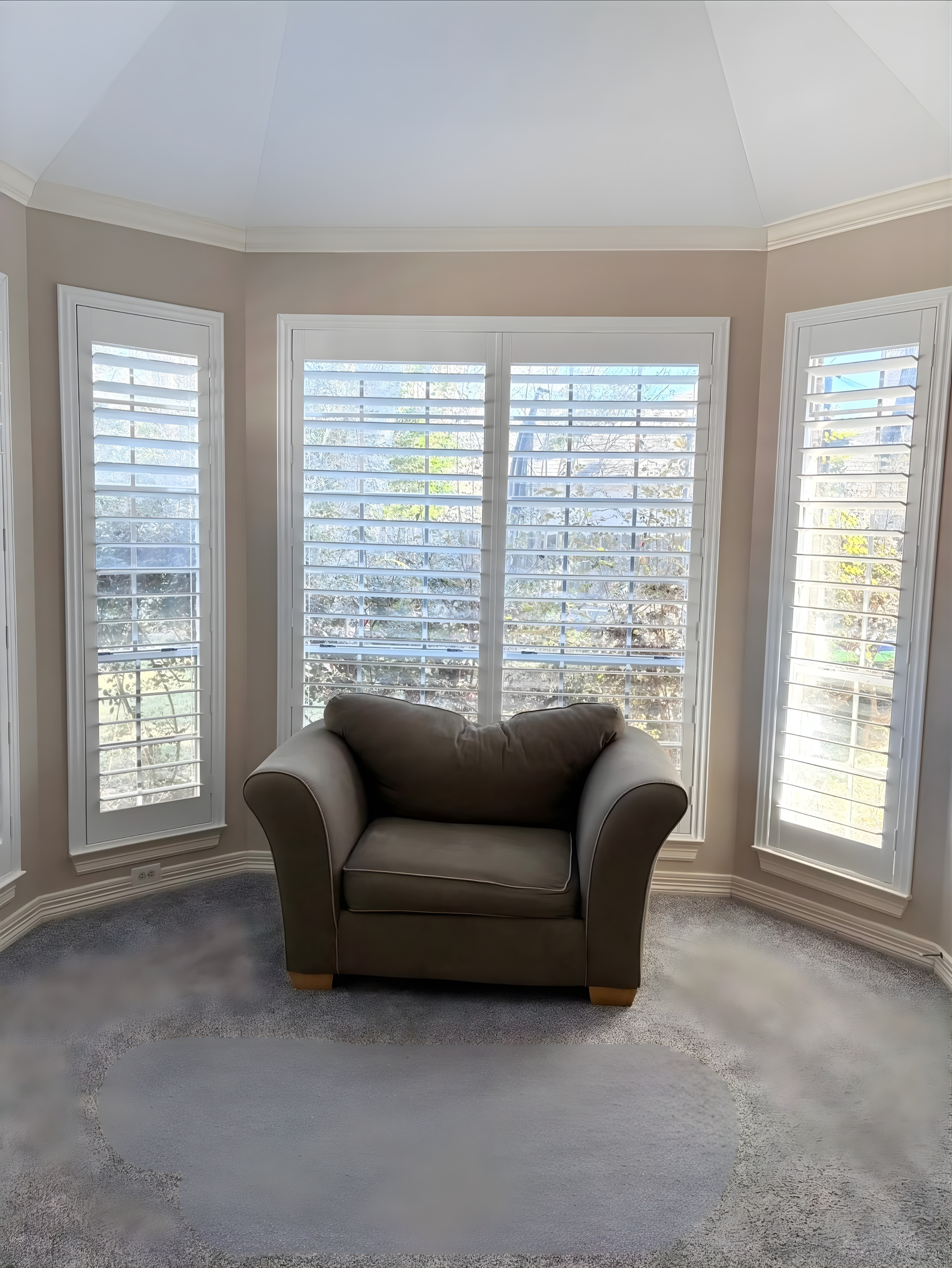 Cozy armchair centered in front of shuttered windows in a light-filled room. Beige walls, floral-patterned carpet.
