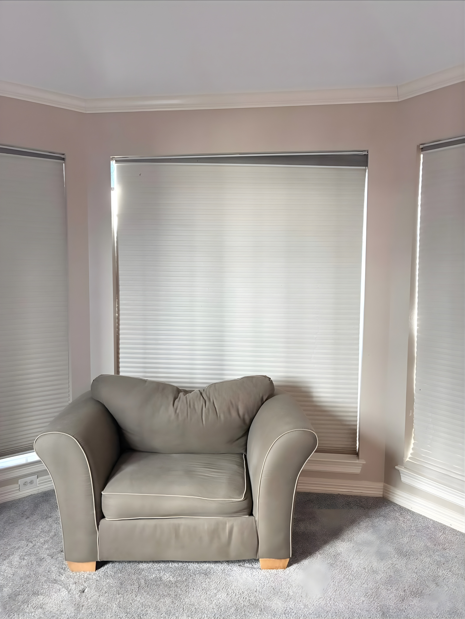 A gray armchair sits in front of a window with white blinds in a room with light pink walls and gray carpet.