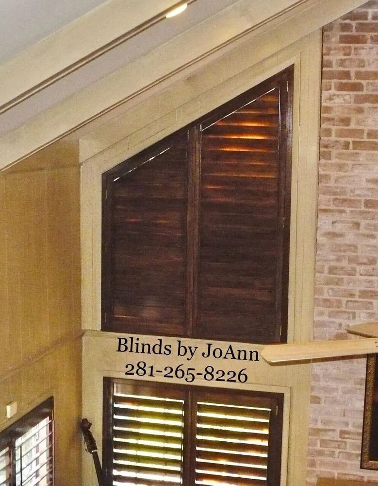 Wooden shutters covering a triangular window, brown finish, set in a light beige frame. Brick wall on the right.